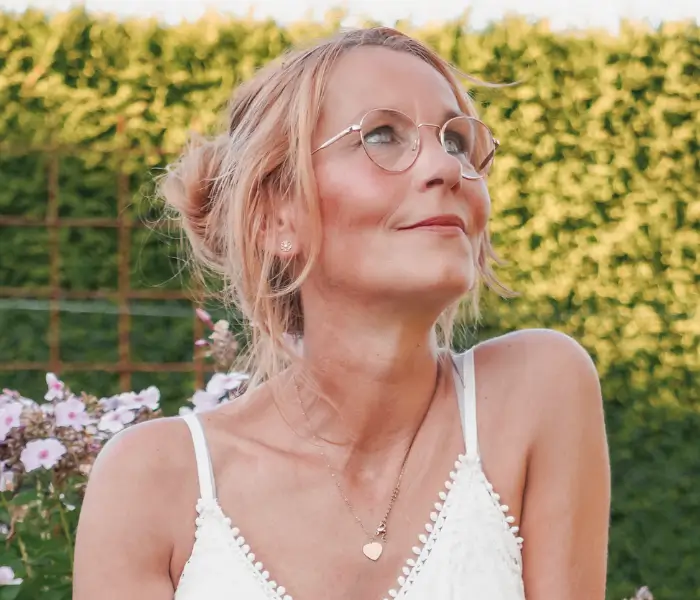 Bettina Jelinek-Stichnoth, psychologische Beraterin, im weißen Sommerkleid im Garten, mit runder Brille und Blick nach oben, umgeben von grüner Hecke und Blüten.