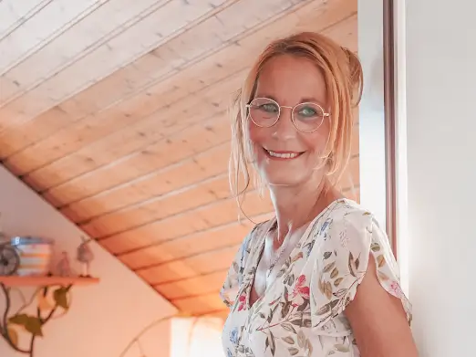 Bettina Jelinek-Stichnoth, psychologische Beraterin, lächelnd im hellen Dachzimmer mit Holzdecke, floralem Kleid und runder Brille, warme und einladende Atmosphäre.