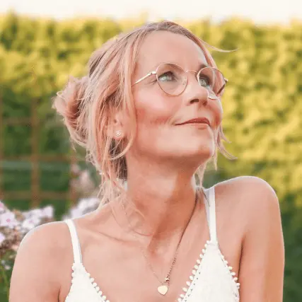 Bettina-Jelinek-Stichnoth-psyhologische-Beraterin-Salzhemmendorf-Startseite Bettina Jelinek-Stichnoth, psychologische Beraterin, im weißen Sommerkleid im Garten, mit runder Brille und Blick nach oben, umgeben von grüner Hecke und Blüten.